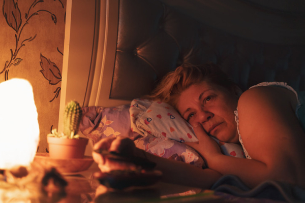 Older woman lying on pillows in bed awake; dim light and a cactus on the bedside table, flowered wallpaper in the background