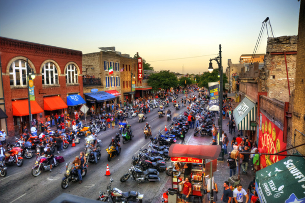 Motorcycle riders and passengers ride up streets of a town in Texas lined with parked motorcycles and rally-goers